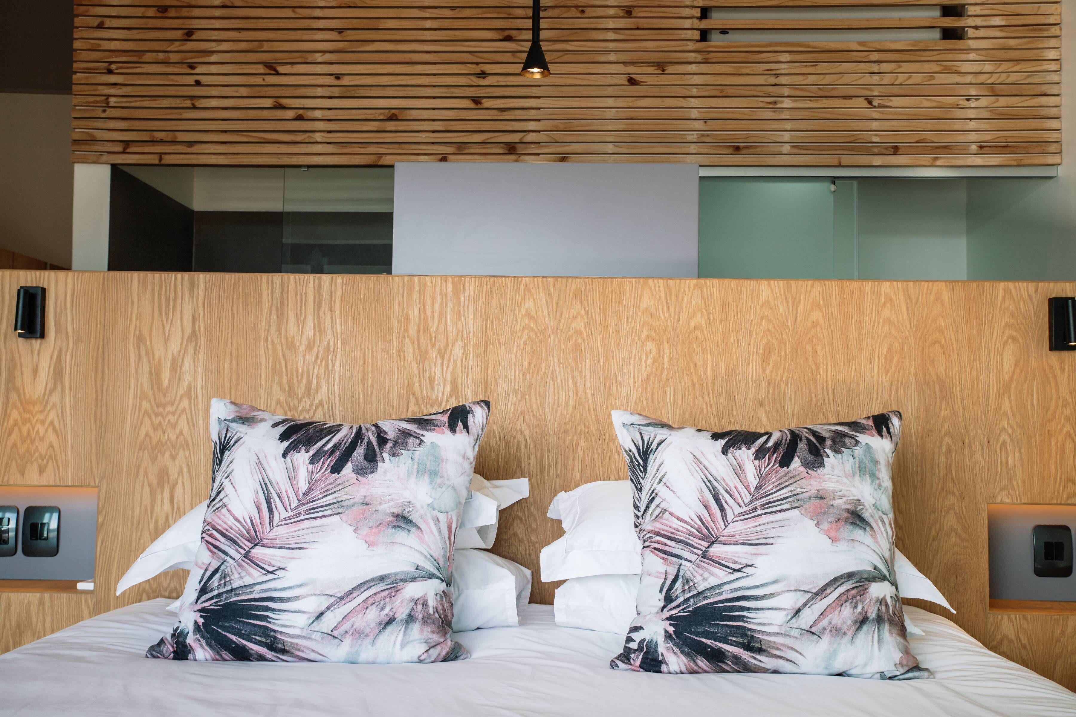 A hotel room with a wooden headboard, two floral pillows, a white bedspread, and a modern pendant light.