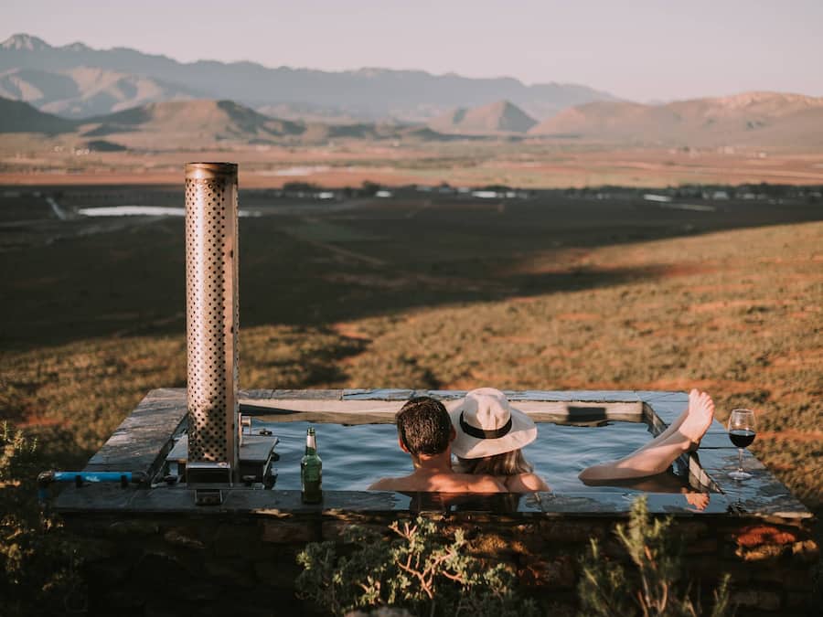 A couple relaxing in a hot tub with a mountain view, a bottle, and a glass of wine.