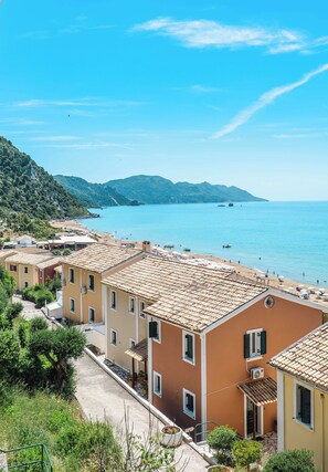 A coastal village with terracotta-roofed buildings, a clear blue sea, and a hilly backdrop.