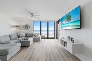 A modern living room with a grey sectional sofa, a flat-screen TV mounted on the wall, and a large window offering a view of the beach. - Sand Dollar 206 Ocean View (St. Augustine)