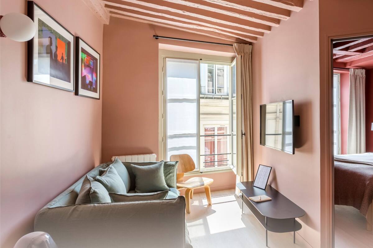 Moderni hotellihuone, jossa on sohva, pieni pöytä kannettavalle tietokoneelle, sängynpäätyyn varustettu sänky ja suuri ikkuna, jossa on verhot.