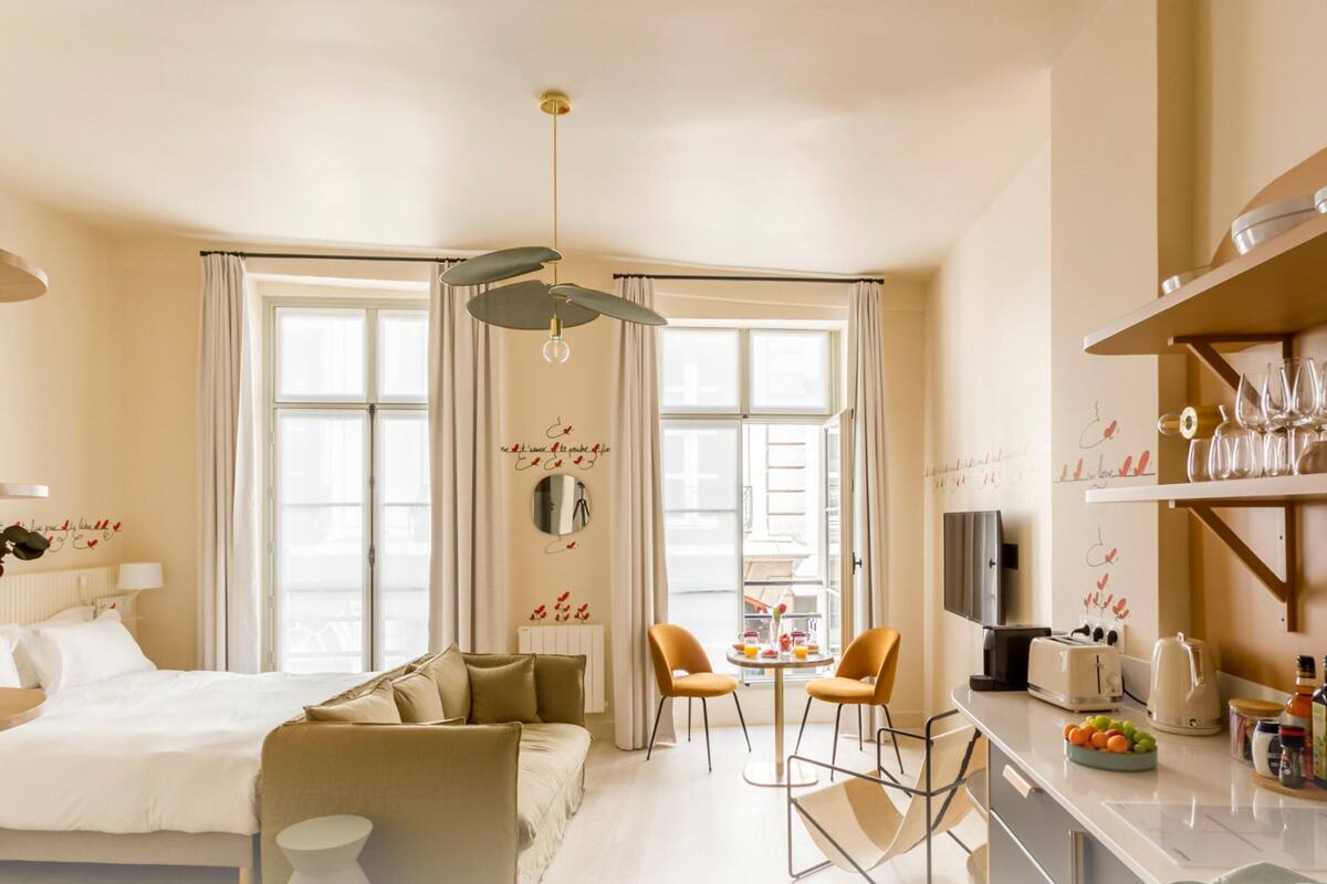 Moderni hotellihuone, jossa on sänky, sohva, ruokailualue tuoleineen, keittiö, jossa on työtaso ja kodinkoneet, sekä suuret ikkunat, joissa on verhot.