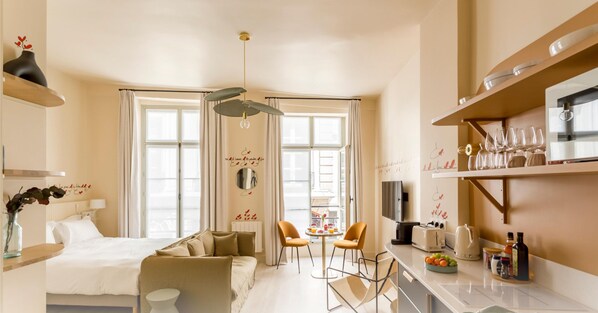 A modern hotel room with a bed, a sofa, a dining area with chairs, a kitchen with a counter and appliances, and large windows with curtains. - Beauquartier - Marais Bourg Tibourg (Paris)