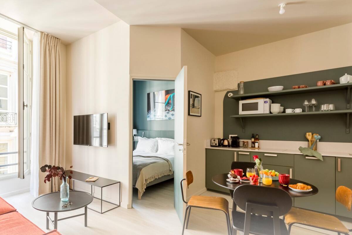 Moderni hotellihuone, jossa on oleskelualue, sänky, pieni keittiö ja näkymä kaupunkiin.