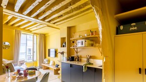 Ein hellgelbes Zimmer mit Holzbalkendecke, einem Essbereich mit Tisch und Stühlen, einer Küche mit Schränken und einer Mikrowelle sowie einem Fenster mit Vorhängen.