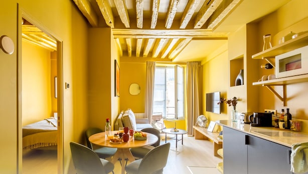 Ein hellgelbes Zimmer mit Holzbalkendecke, einem Esstisch mit Stühlen, einer Couch, einem kleinen runden Tisch und einer Küchenzeile mit Spüle und Arbeitsplatte.