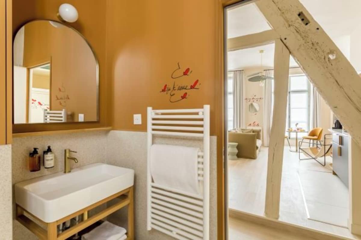 Ein modernes Badezimmer mit einem weißen Waschbecken, einem Handtuchhalter und einem Spiegel. Ein Fenster bietet Blick auf einen Wohnbereich.