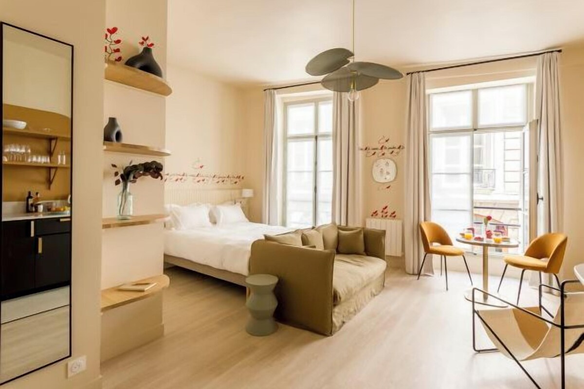Une chambre moderne avec un lit, un canapé et des fauteuils, un coin cuisine et de grandes fenêtres.