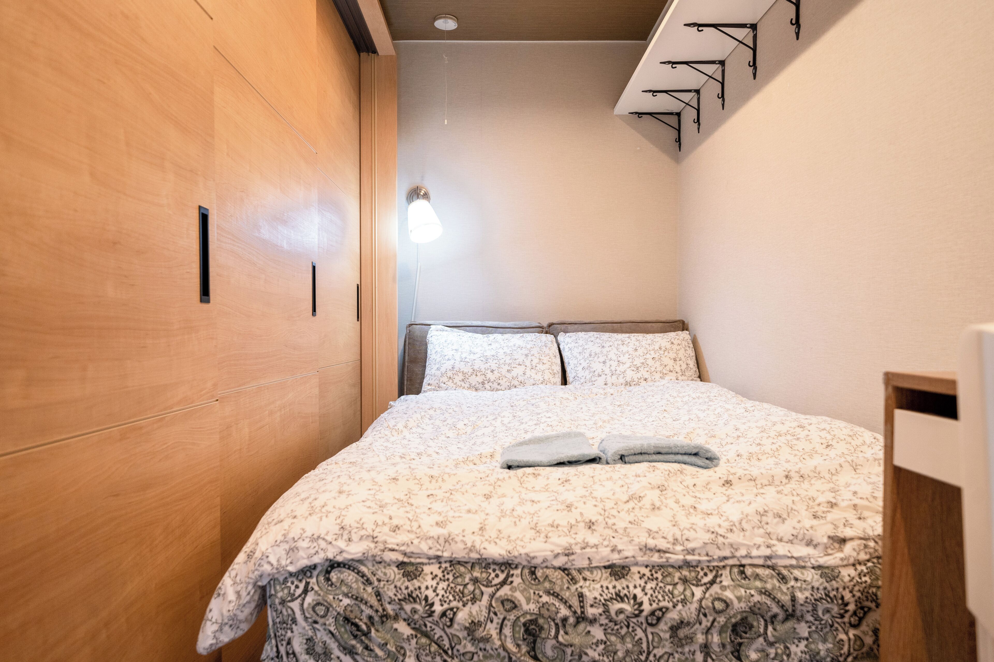 A bedroom with a wooden wardrobe, a bed with floral bedding, and a wall-mounted light fixture.