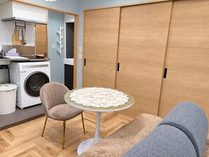 Een ruimtelijk ingerichte leefruimte met een wasmachine, een eettafel met een tafelkleed met bloemenprint, een comfortabele stoel en een kledingkast.