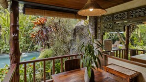 Une terrasse en bois avec une table et des chaises, entourée d’une végétation tropicale luxuriante et agrémentée d’un petit élément décoratif aquatique.