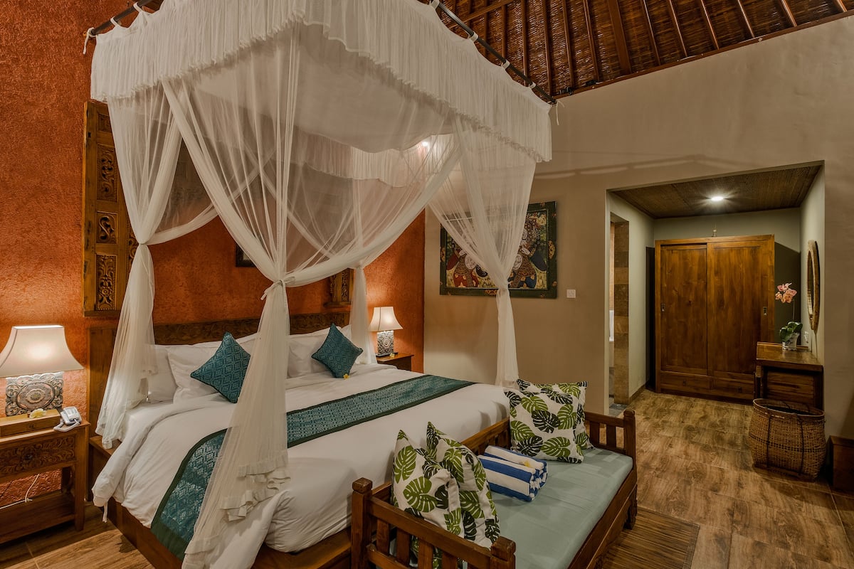 A four-poster canopy bed with white linens and teal pillows, a wooden nightstand with a lamp, and a wooden bench with a green cushion.
