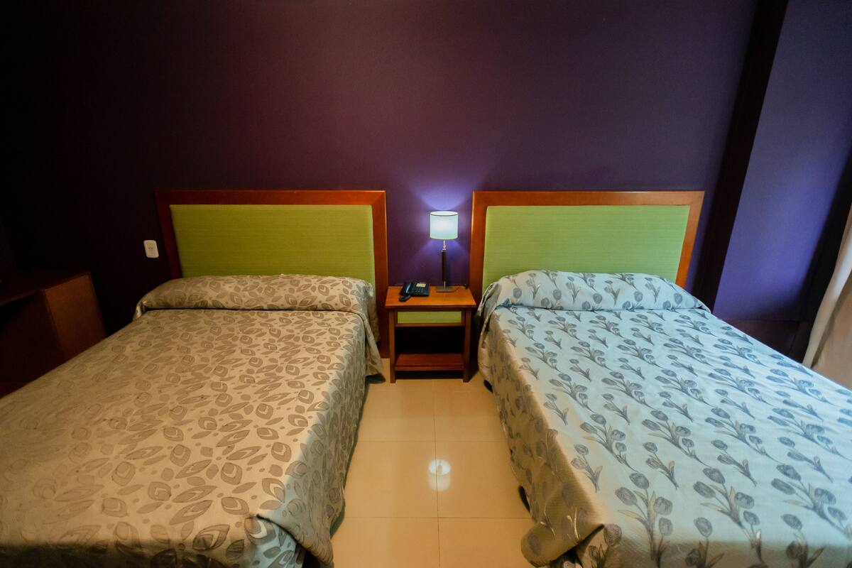 A hotel room with two beds, a nightstand with a lamp, and a small table.