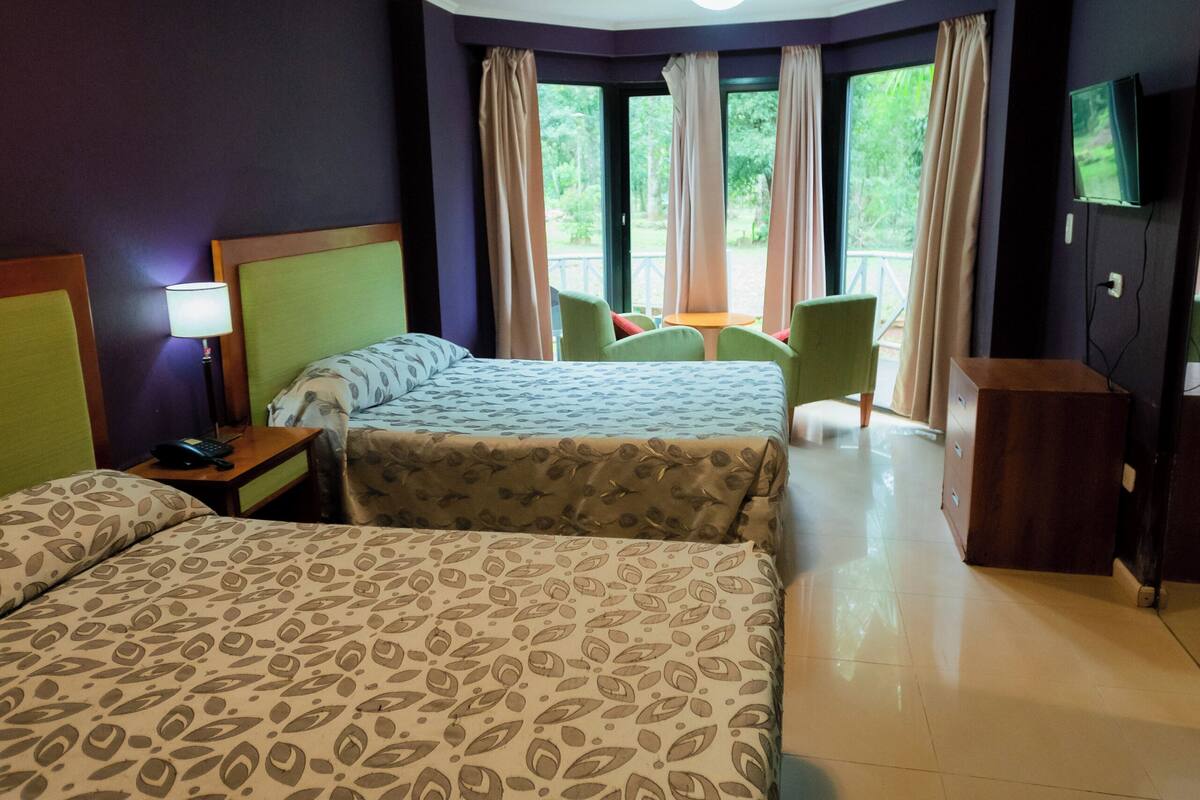 A hotel room with two beds, a desk, a chair, a TV, and a balcony with curtains.