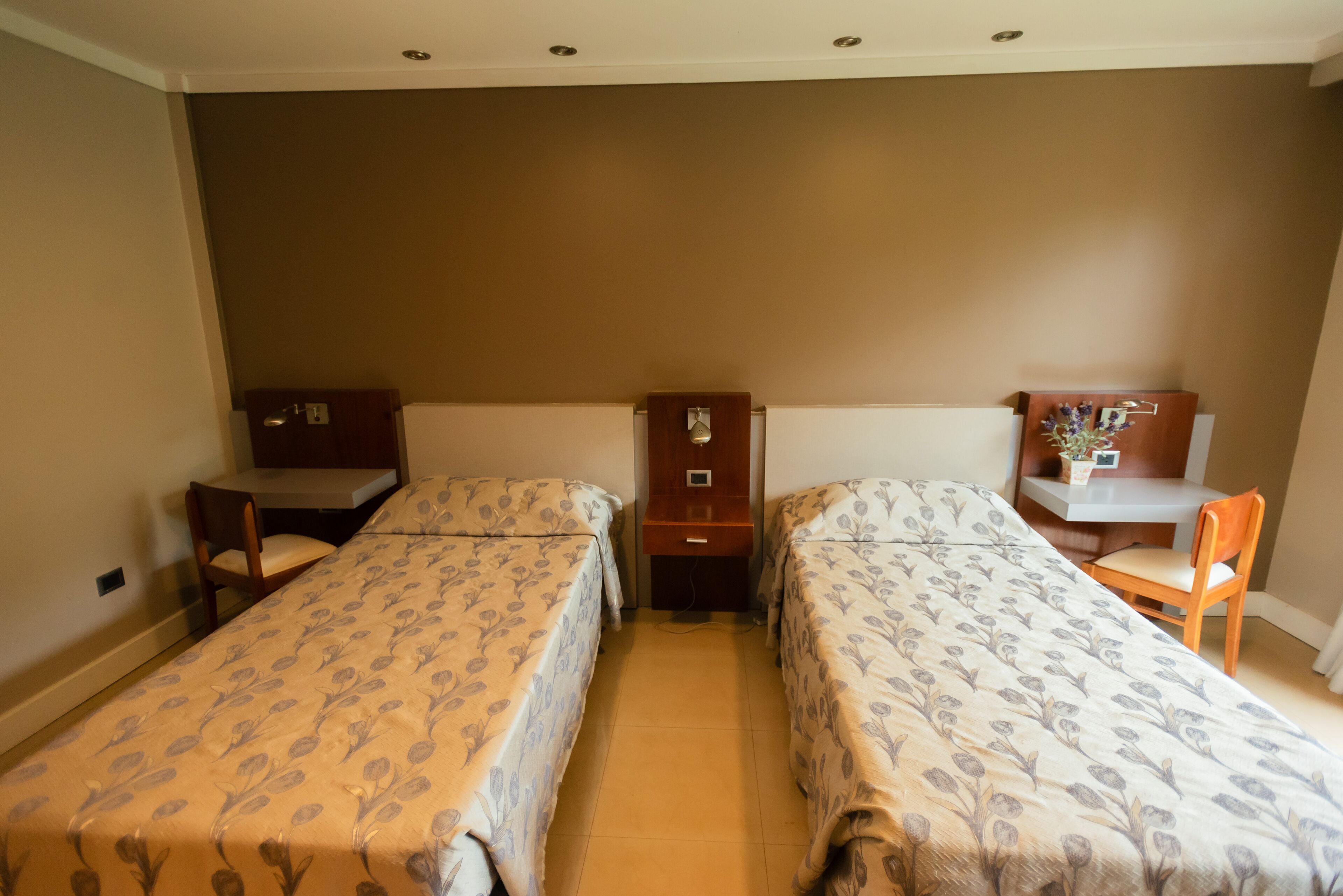 A hotel room with two single beds, a desk, a chair, and a small table with a vase.