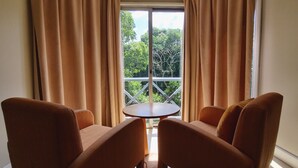Une pièce avec deux fauteuils marron et une petite table ronde, une grande fenêtre avec des rideaux, et une vue sur des arbres et un balcon.