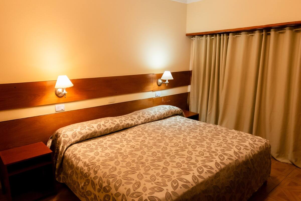 A hotel room with a large bed, wooden headboard, two bedside lamps, and a curtain.