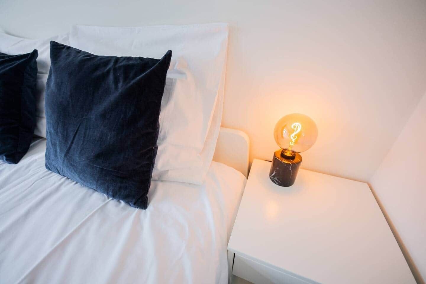 Ein ordentlich bezogenes Bett mit weißen und dunkelblauen Kissen, ein weißer Nachttisch mit einer beleuchteten Globenlampe und eine weiße Wand.