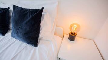 Ein ordentlich bezogenes Bett mit weißen und dunkelblauen Kissen, ein weißer Nachttisch mit einer beleuchteten Globenlampe und eine weiße Wand.