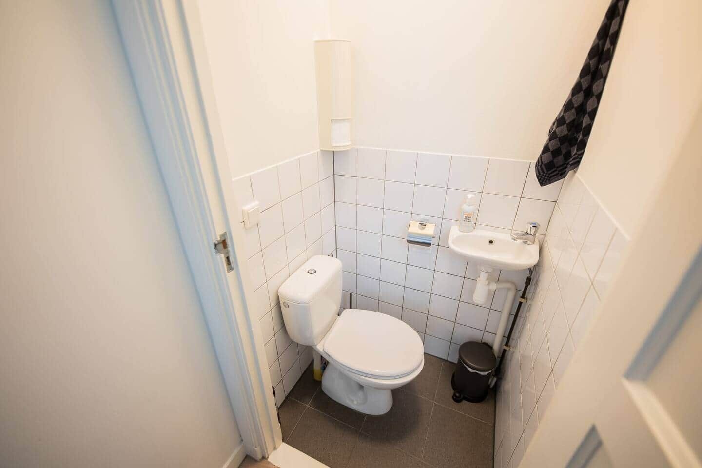 Un bany petit i net amb rajoles blanques, un vàter, un lavabo i un gresol.