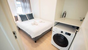 Une chambre d’hôtel de taille réduite, équipée d’un lit, d’une machine à laver et d’une petite étagère avec un miroir.