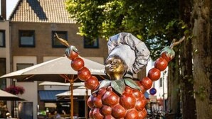 Une statue représentant une figure coiffée d’un chapeau de chef et dont les bras sont faits de sphères rouges, dressée sur un piédestal dans un espace extérieur aménagé avec des tables et des parasols.