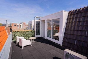 Terraza en la azotea con pared blanca, puerta de cristal, una mesita, dos sillas blancas y un ventilador.