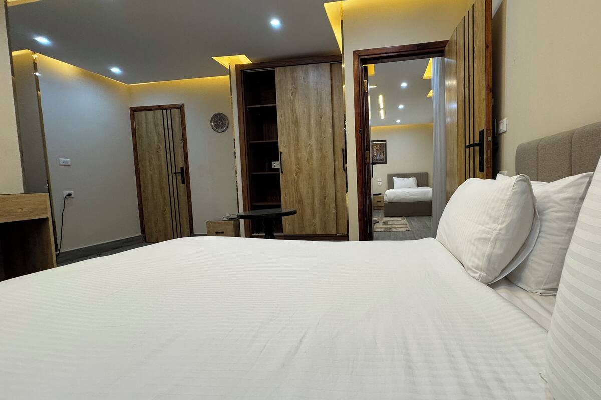 A hotel room with a large bed, a wardrobe, a desk, and a door leading to another room.