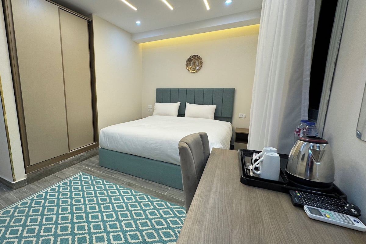 A hotel room with a bed, a desk with a kettle, a coffee maker, and a remote control.