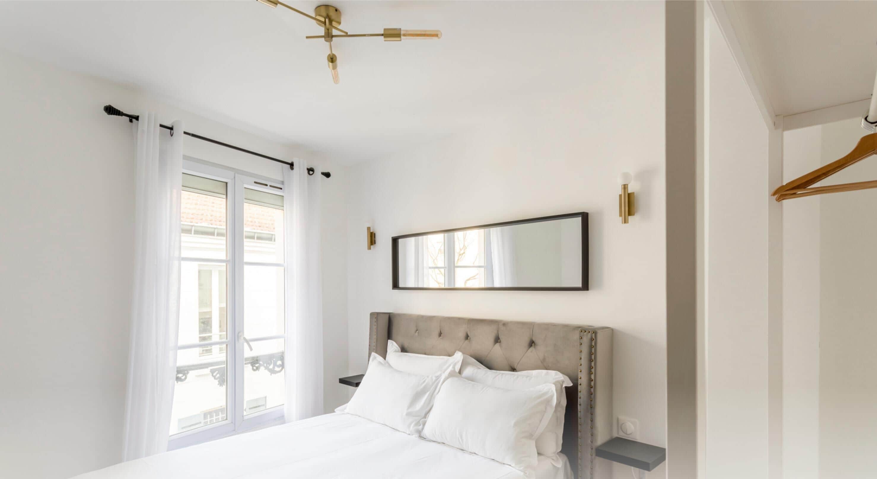 A bedroom with a large bed, a ceiling fan, a window with curtains, and a wall-mounted mirror.