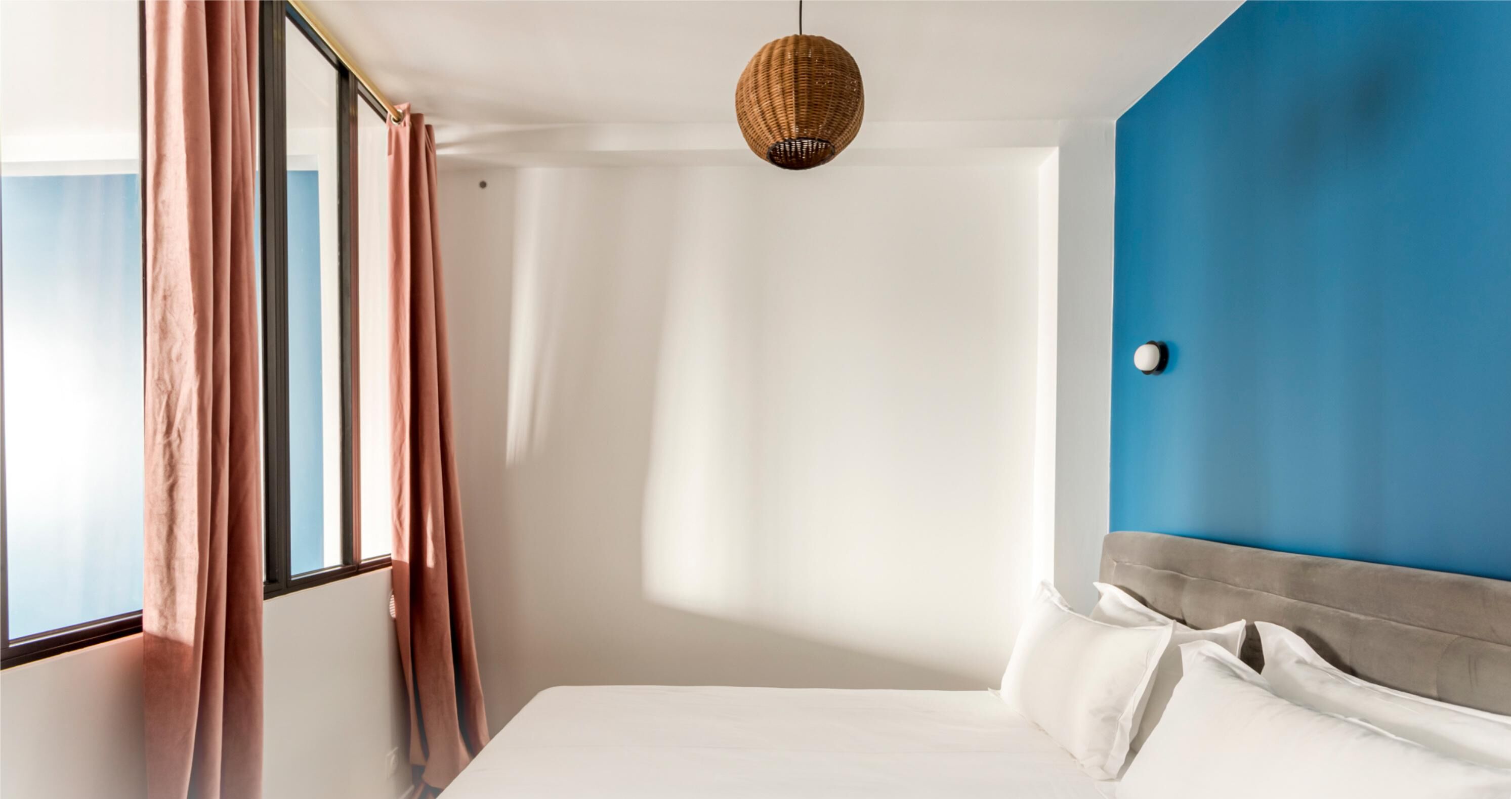 A hotel room with a large window, a bed with white linens, a blue accent wall, and a woven pendant light.