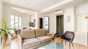 Una sala de estar moderna con un sofá beige, una mesa de centro de vidrio y una silla negra. Se observa un comedor al fondo, una cocina con baldosas estampadas y un vestidor con espejo.