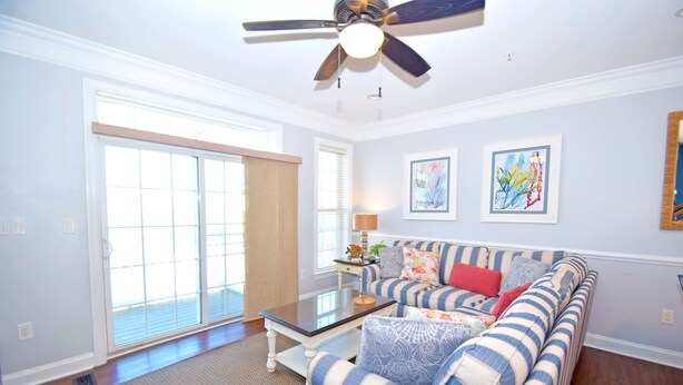A living room with a sofa, coffee table, and ceiling fan. There are two windows and a door.