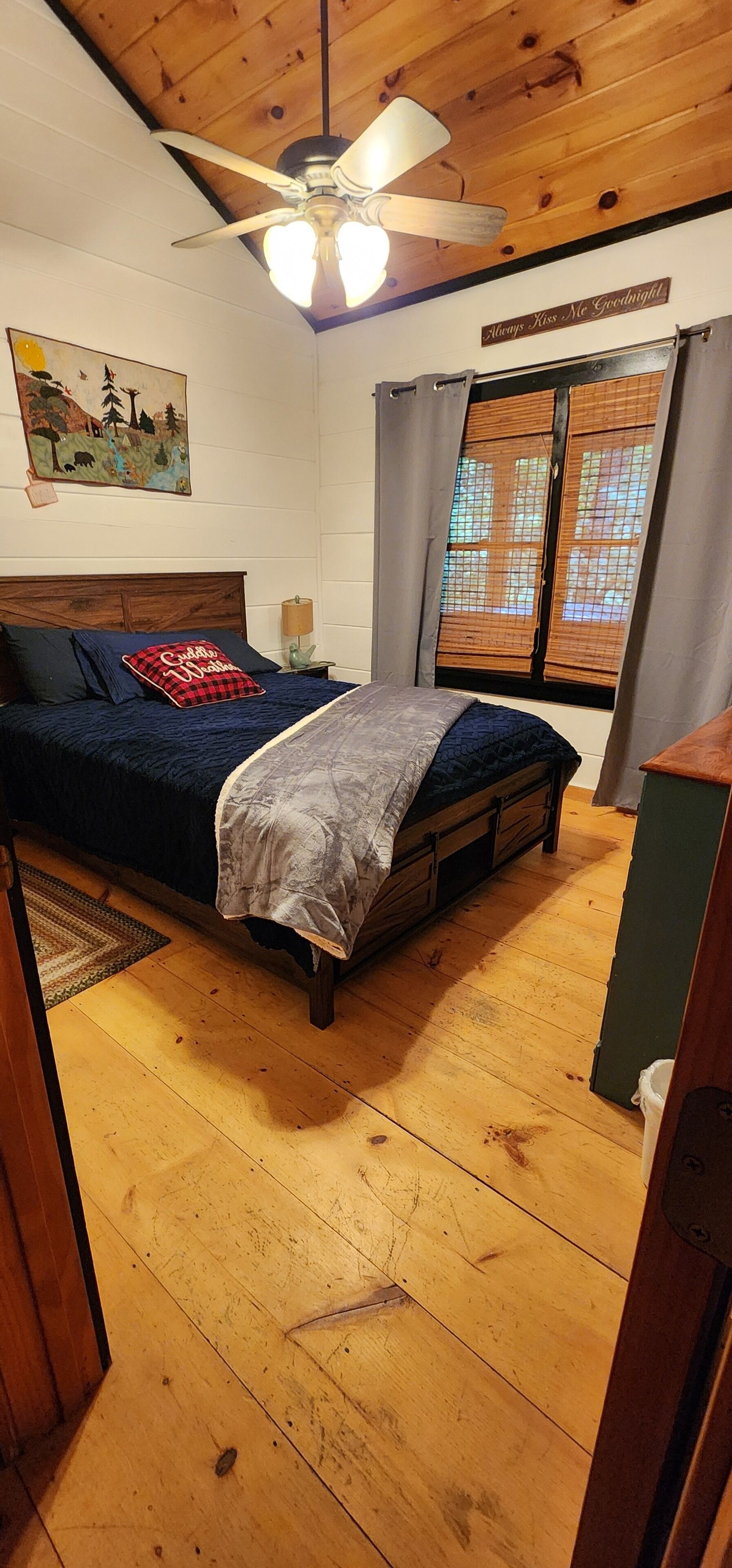 Un dormitorio con techo de madera, una cama con cubrecama azul y blanco, un ventilador de techo, una mesita de noche con lámpara, una ventana con persianas de bambú y un cuadro en la pared.