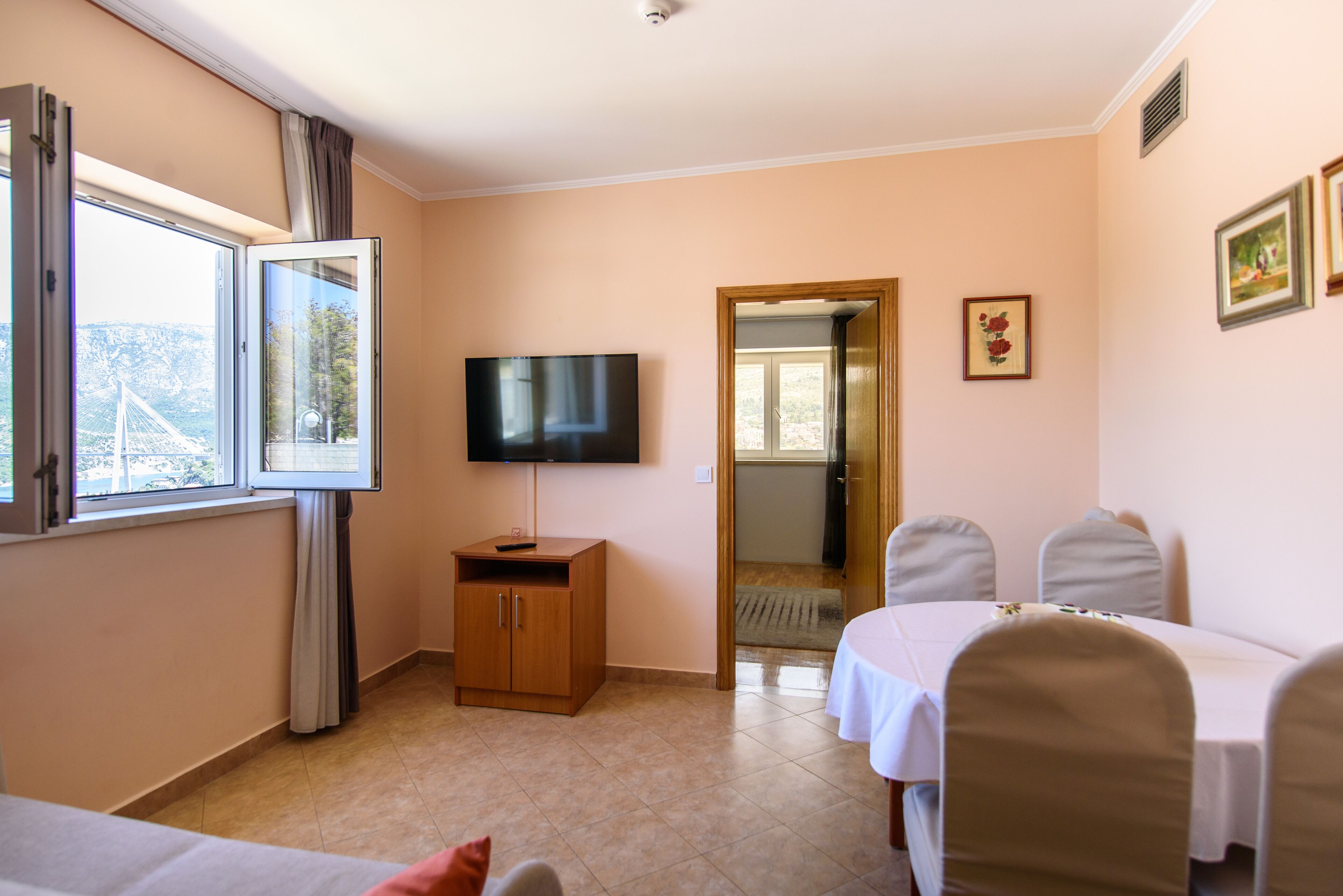 Een hotelkamer met een televisie, een houten kast, een eettafel met stoelen en een raam met gordijnen.