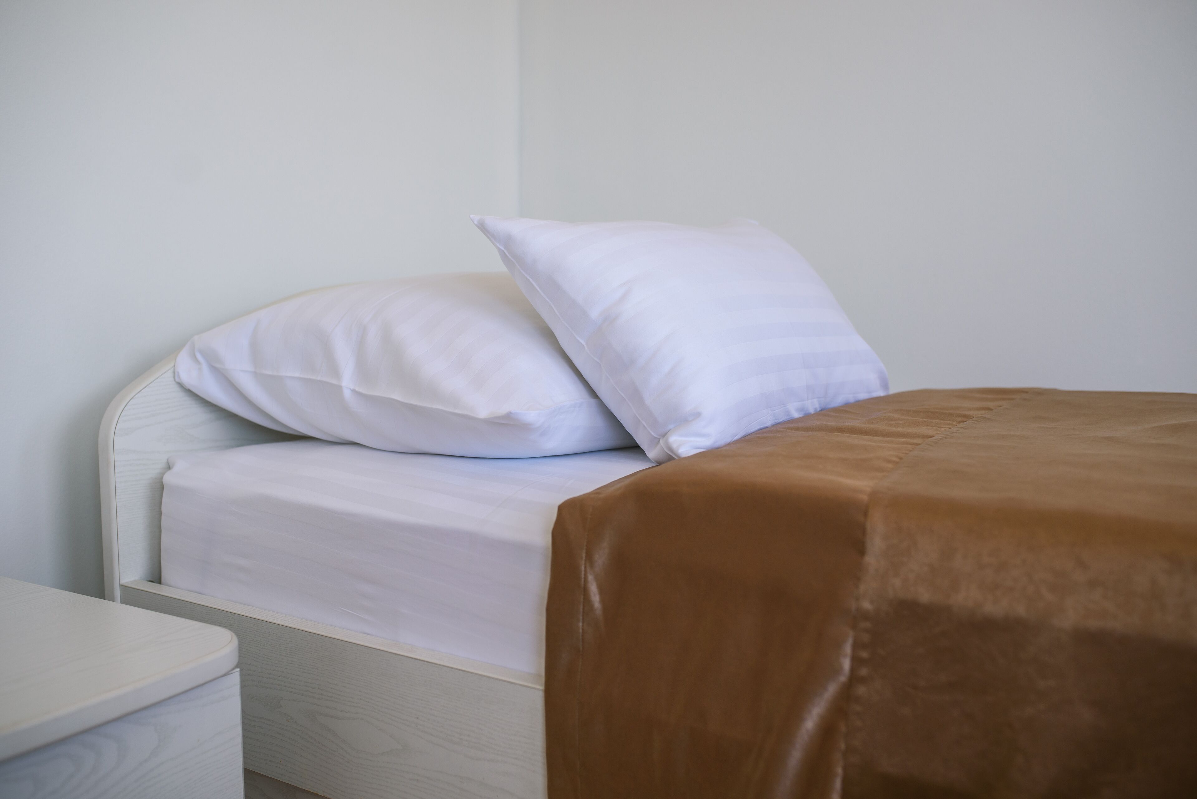 A bed with white bedding and a brown blanket, a white nightstand, and a plain wall.