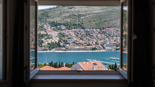 A coastal cityscape viewed from a window, featuring a mix of residential and commercial buildings, a marina, and a hilly backdrop.
