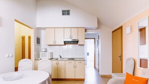 A compact kitchen with white cabinets, a microwave, and a sink. There is a dining table with a white tablecloth and a glass bowl. A bright orange pillow is on a white sofa.