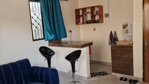 Una sala de estar con un sofá azul, una mesa de centro blanca, un taburete de bar amarillo y blanco, un ventilador de techo y una zona de cocina con mostrador y estanterías.