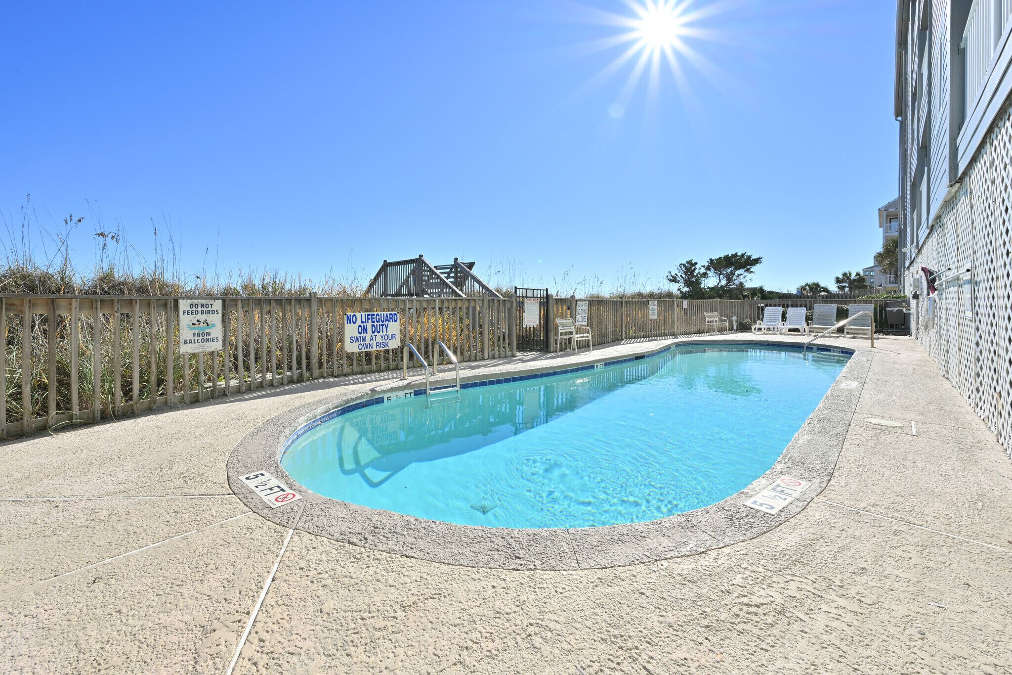 A swimming pool with a wooden fence, signs indicating no lifeguard on duty, and a clear blue sky.