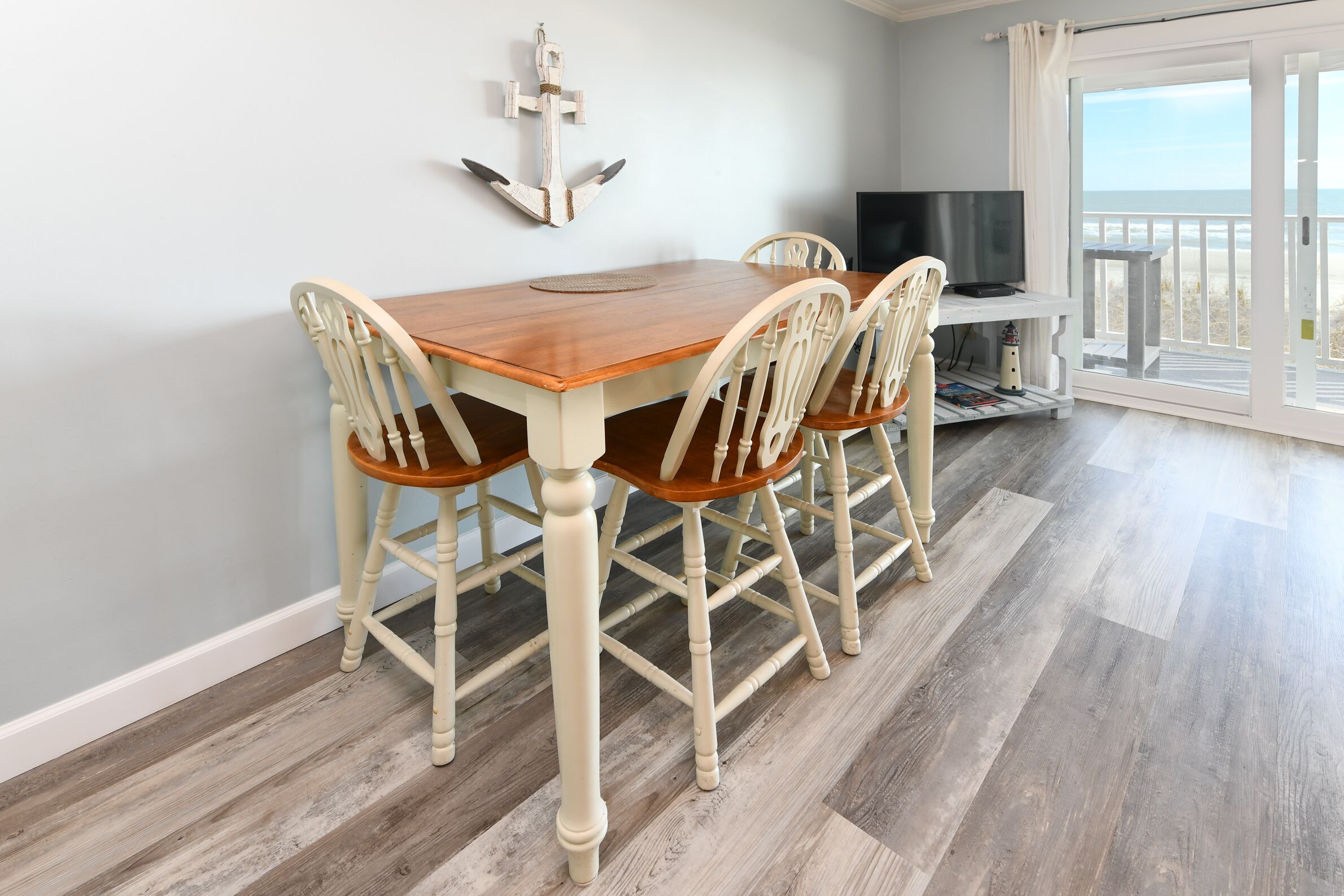 A dining area with a wooden table and four chairs, a television, and a view of the beach through a glass door.