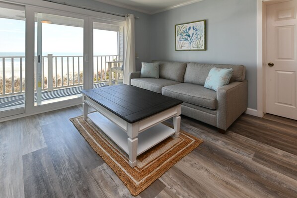 A room with a sliding glass door leading to a balcony, a grey sofa with star-patterned cushions, a wooden coffee table, and a framed artwork on the wall.