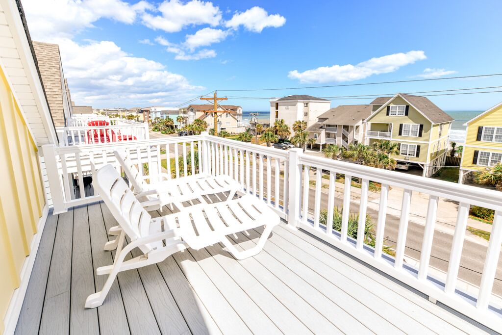 Une terrasse en bois avec des chaises blanches et une balustrade, donnant sur une plage bordée de maisons et sous un ciel dégagé.