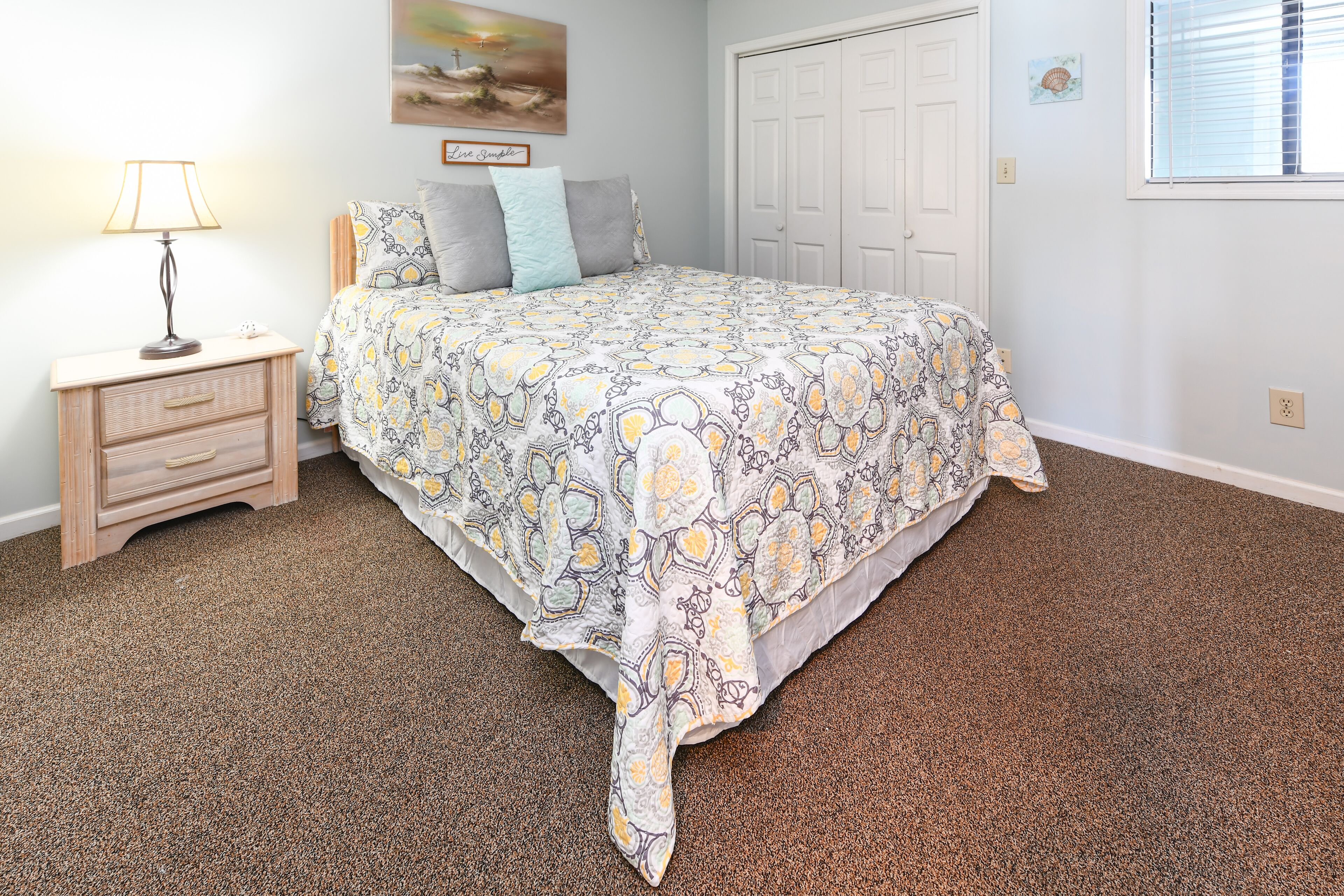 A bedroom with a patterned bedspread, a bedside table with a lamp, a framed picture on the wall, and a carpeted floor.