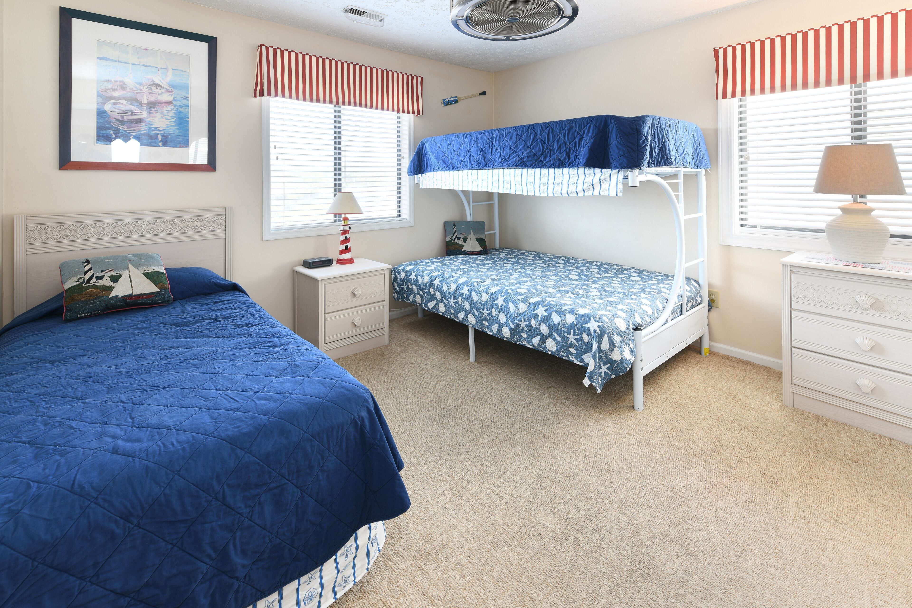 Een slaapkamer met twee bedden, een stapelbed, een nachtkastje met lamp, een dressoir en een lijst met een afbeelding aan de muur.
