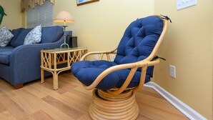 Una habitación con una silla azul con cojines acolchados y estructura de madera, un sofá azul con cojines estampados y una pequeña mesita de madera con una lámpara.