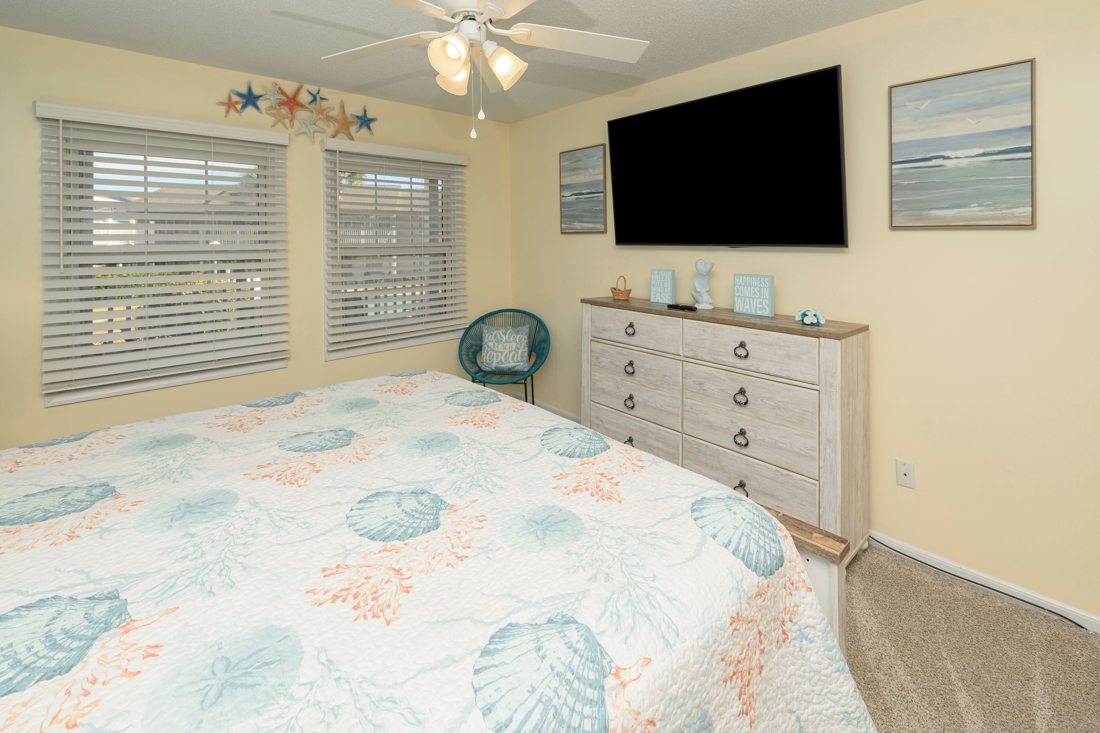 Un dormitorio con cama, cómoda, silla, ventilador de techo y dos ventanas con persianas.