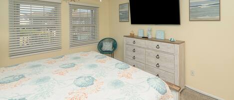 Un dormitorio con cama, cómoda, silla, ventilador de techo y dos ventanas con persianas.