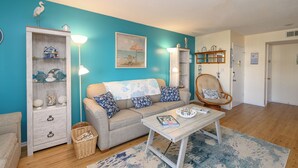 A living room with a teal wall, a sofa with patterned cushions, a wooden coffee table, and a wicker basket. - 2nd Row 3 BR Condo with a Shared Pool! Floral Villas D13  (Surfside Beach)