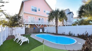 Ein zweistöckiges Haus mit rosa Fassade, ein Garten mit Swimmingpool und ein weißer Zaun.